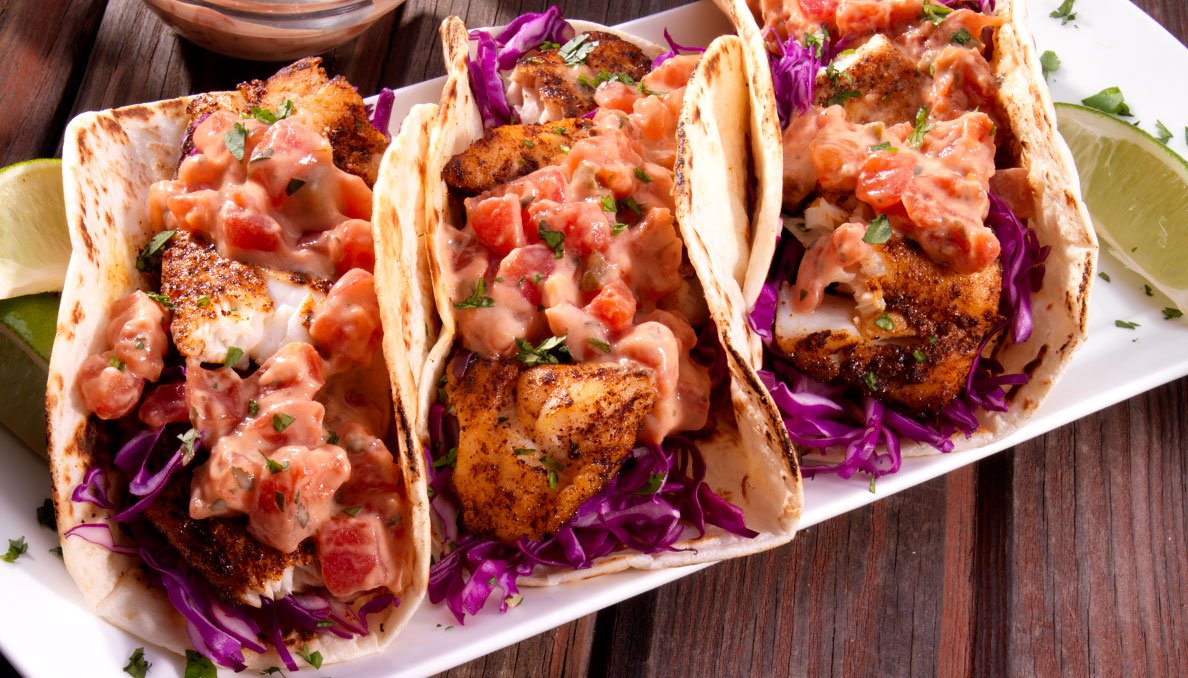 Image of three fish tacos topped with a lime and cilantro aioli  with purple cabbage on a white plate