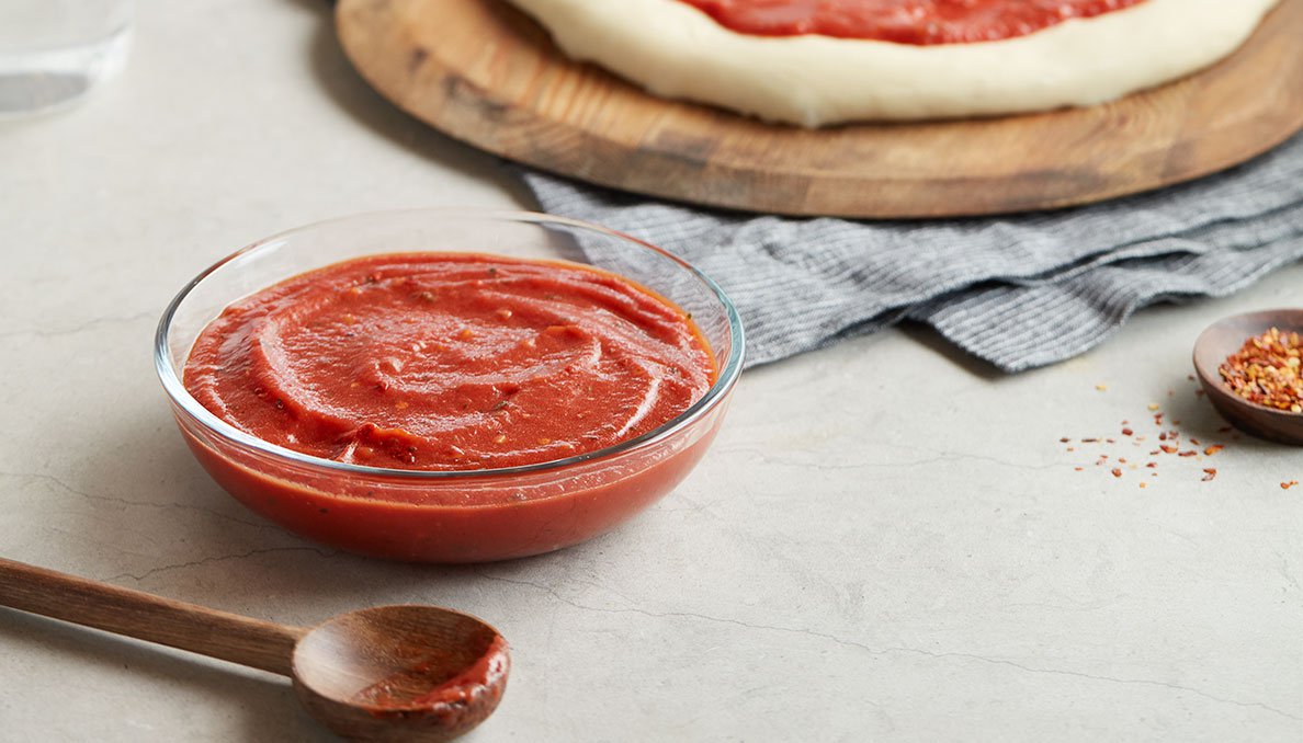 Image of easy no cook pizza sauce in a clear glass bowl with wooden sppon and pizza dough in background