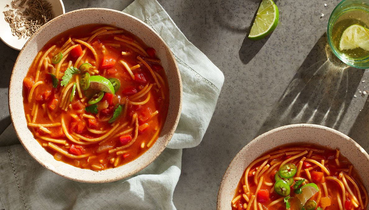 Overhead image of fideo soup with tomato base diced tomatoes noodles jalapeno slices and lime wedges
