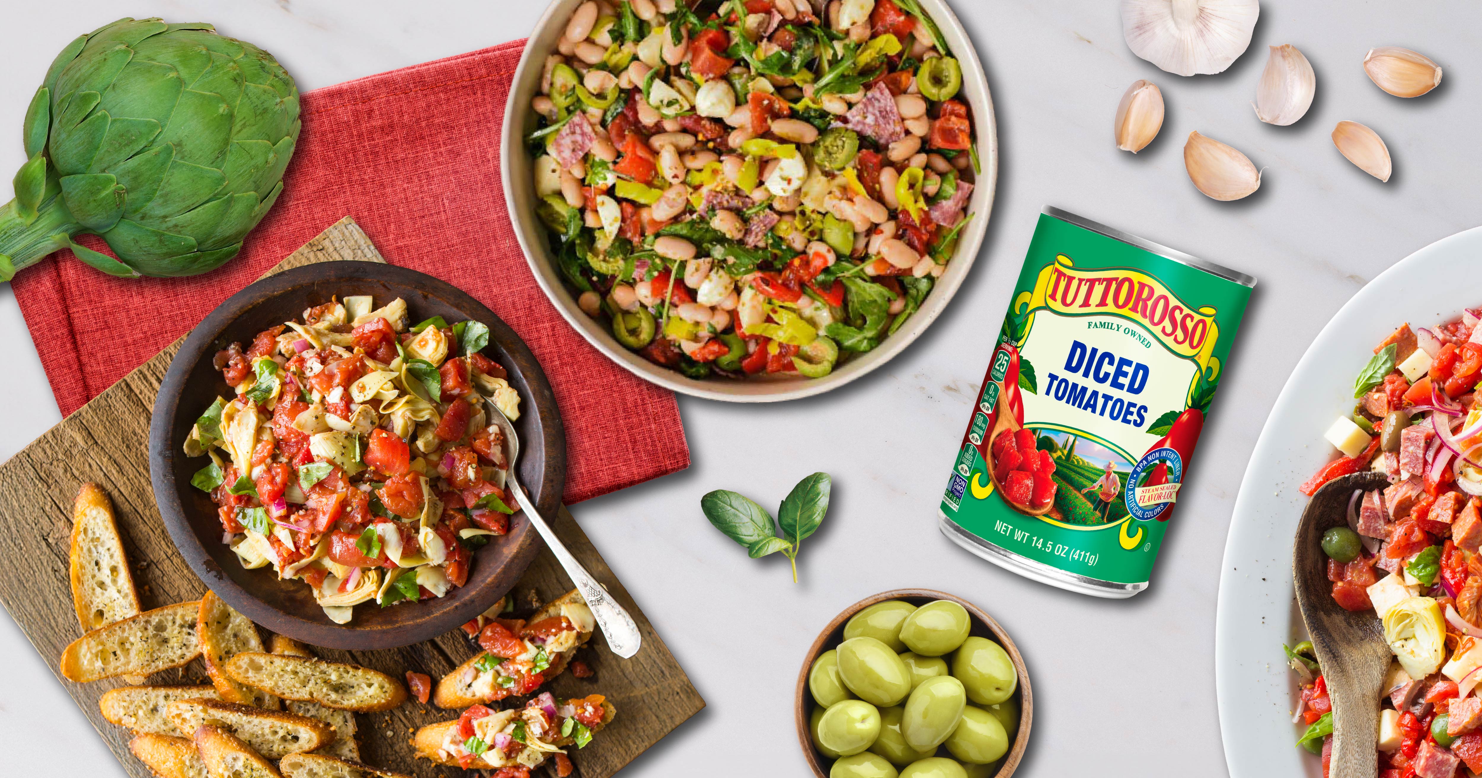 Overhead food scene featuring Mediterranean-style dishes made with diced tomatoes: a bowl of bruschetta with toasted bread, a white bean and vegetable salad, and an antipasto-style salad with olives and cheese. Fresh ingredients like an artichoke, garlic cloves, basil leaves, and green olives are arranged around a can of Tuttorosso diced tomatoes on a light countertop.